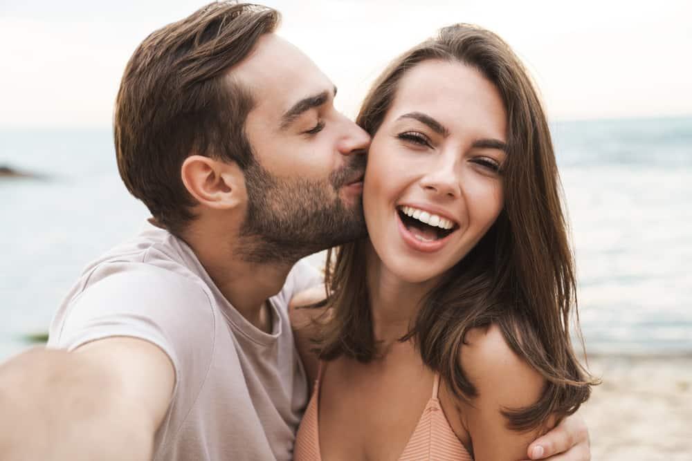 young happy man kissing and hugging beautiful woman while taking selfie photo on sunny beach