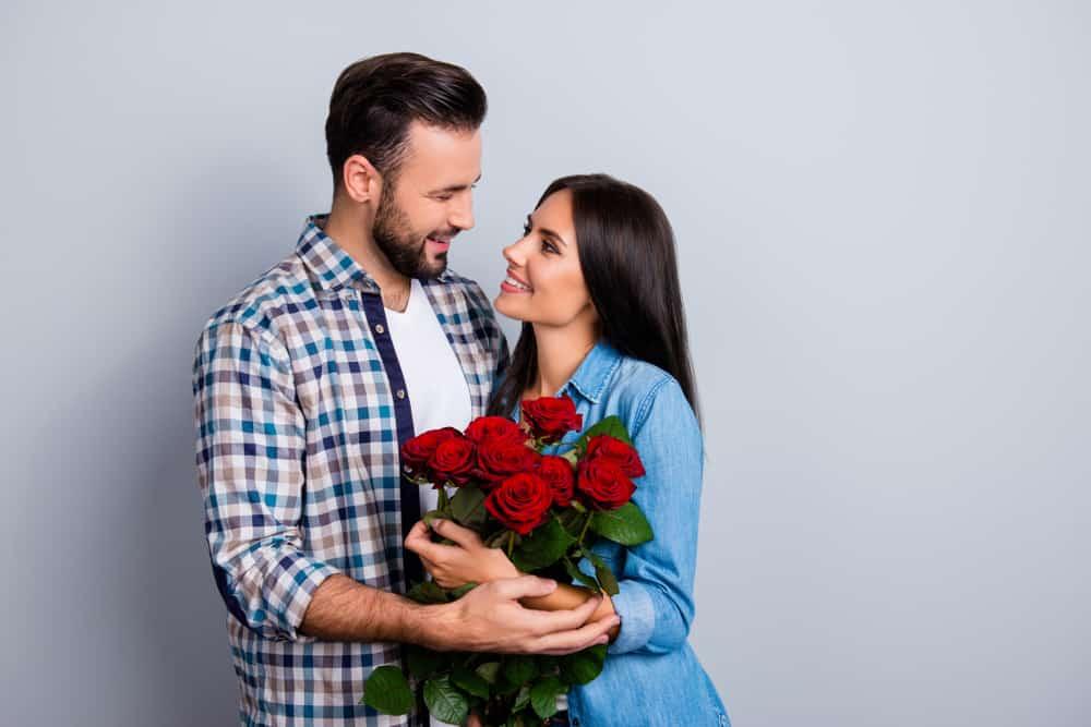 Beautiful, happy, positive couple embracing, looking to each other, holding bouquet of red roses