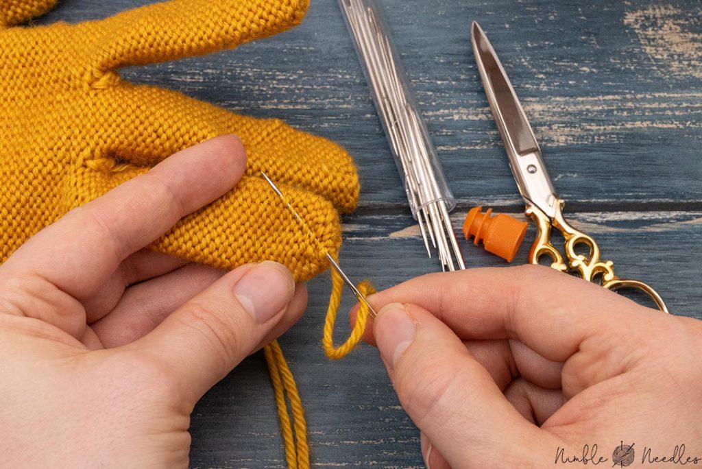 someone weaving in the tails of the knitted glove with a sharp tapestry needle going diagonally