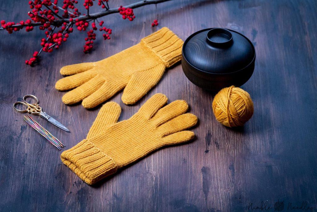 a pair yellow knitted gloves with fingers on a wooden board with knitting tools in the background