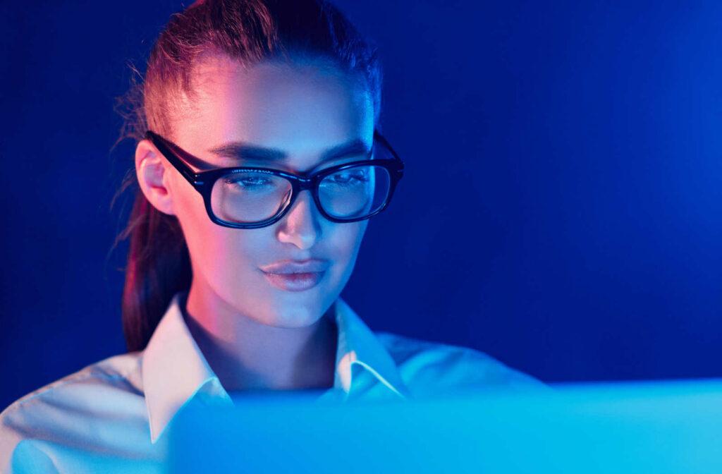 A woman looking at a laptop with the blue light radiating off of her face and glasses.