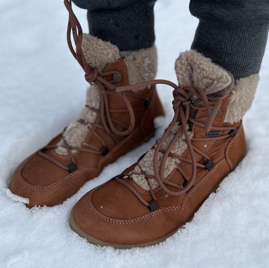 A close up side view of a pair of feet standing in snow wearing Be Lenka Bliss barefoot winter boots in brown