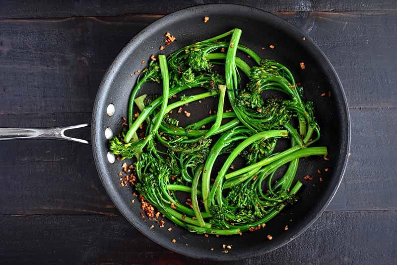 A close up top-down picture of a black frying pan with stir fried broccolini in garlic, set on a dark wooden surface.