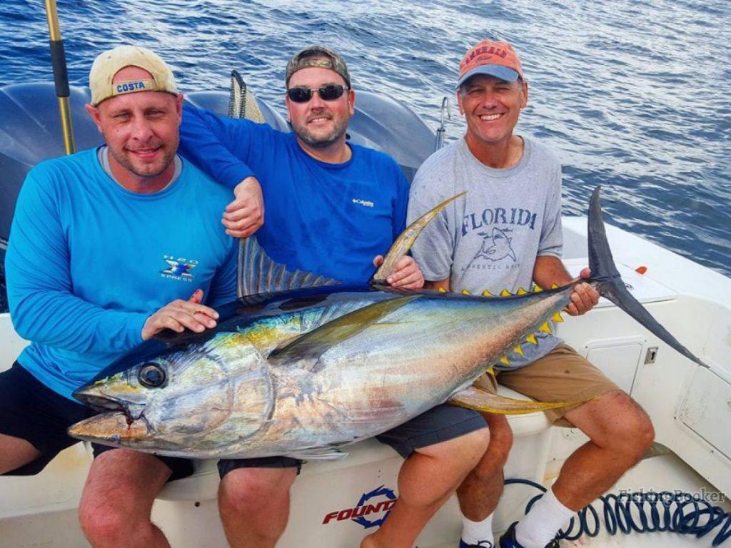 A smiling fisherman standing on a boat in a cap and sunglasses, holding a big Bluefin Tuna, with murky waters behind him