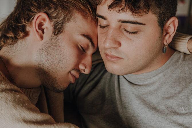 Casual Laughing Gay Couple Toasting Glasses of Red Wine in a Park Picnic Sitting on Green Grass