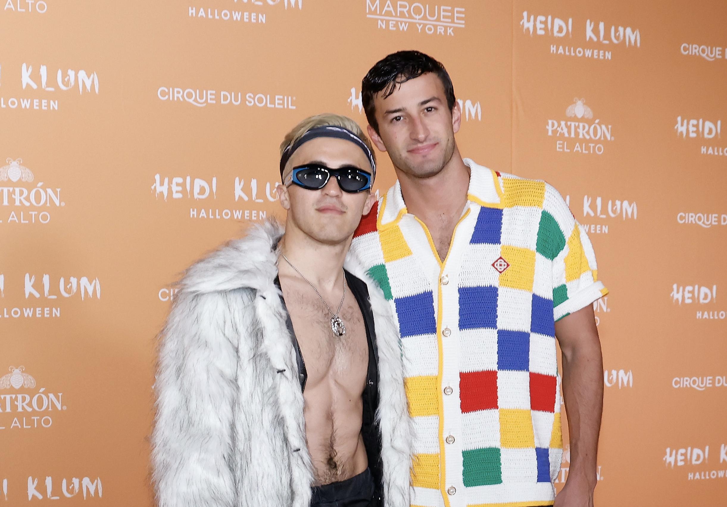 Chris Olsen, wearing a furry coat open to reveal a ripped chest, a necklace, black sunglasses, and a bandana wrapped around his bleached hair stands next to Patrick Johnson. Johnson smiles softly in a red, blue, yellow, and green checkerboard white t-shirt like Allan from