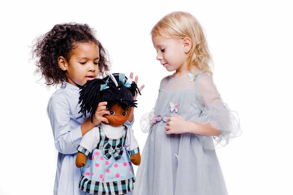 Little fashionable girls are friends of two different nationalities. African American girl showing doll to her friend