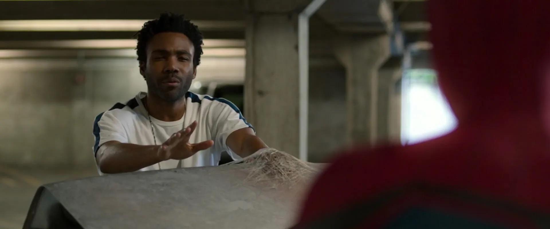 Donald Glover as Aaron Davis in Spider-Man: Homecoming. He’s got one hand webbed to the top of a car’s trunk; Spider-Man appears blurrily in the foreground.
