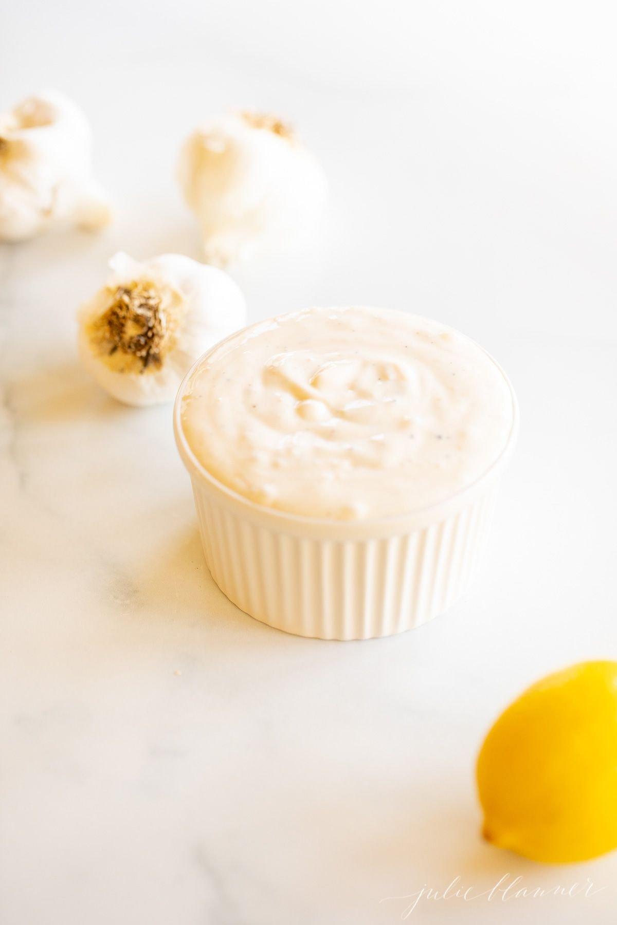 On a marble surface, a white ramekin dish full of fresh garlic aioli, with a head of garlic to the side.