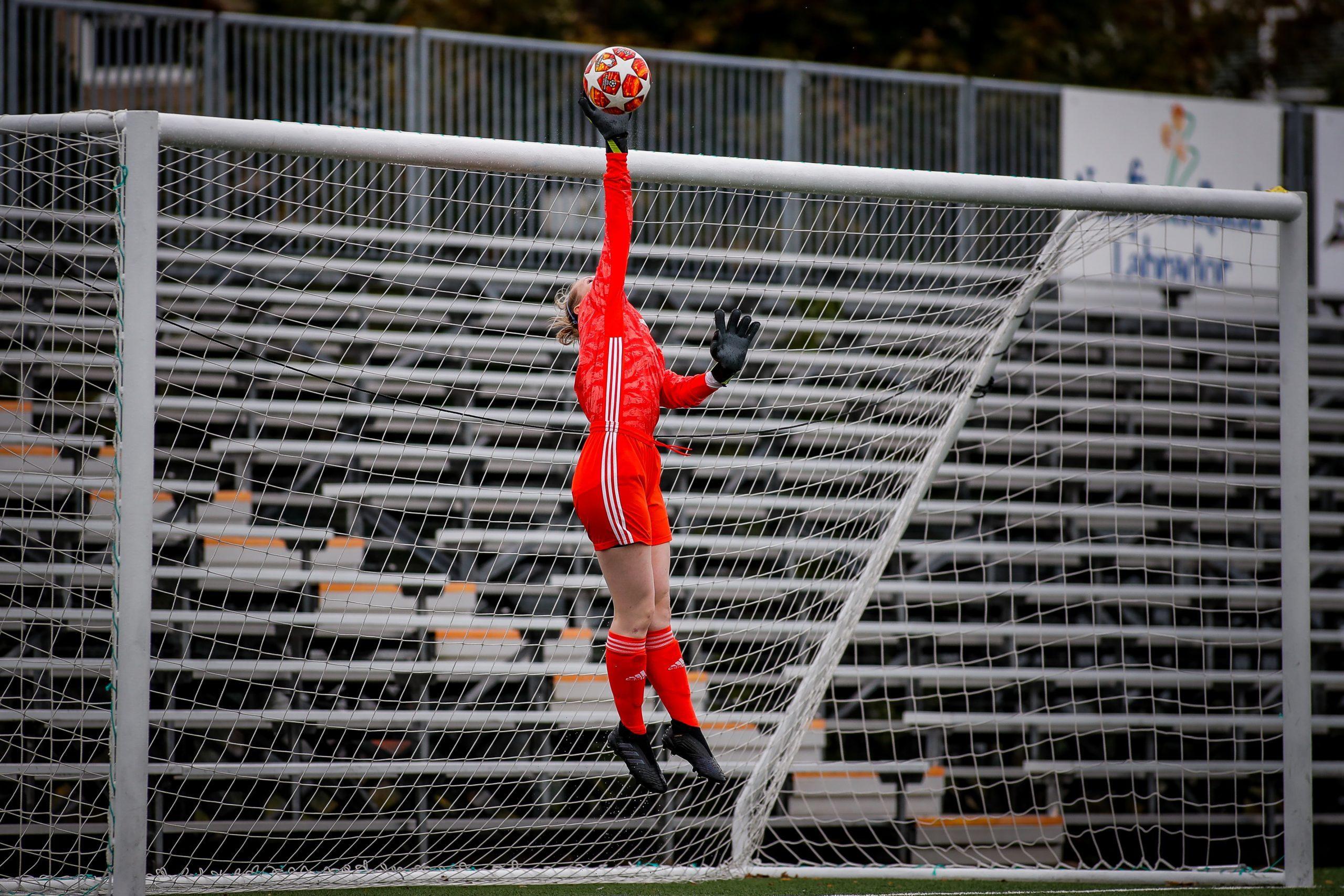 A female goalkeeper in red leaps to tip the ball over the crossbar in the rain