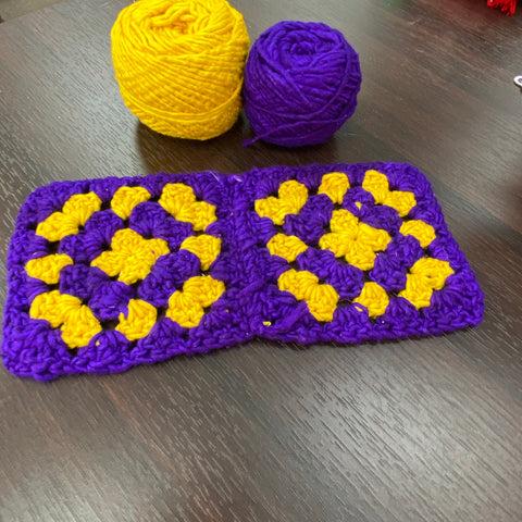 Two granny squares, made from purple and yellow worsted weight yarn are laying on a dark wooden surface. Behind them are the two cakes of yellow and purple worsted weight silk yarn.
