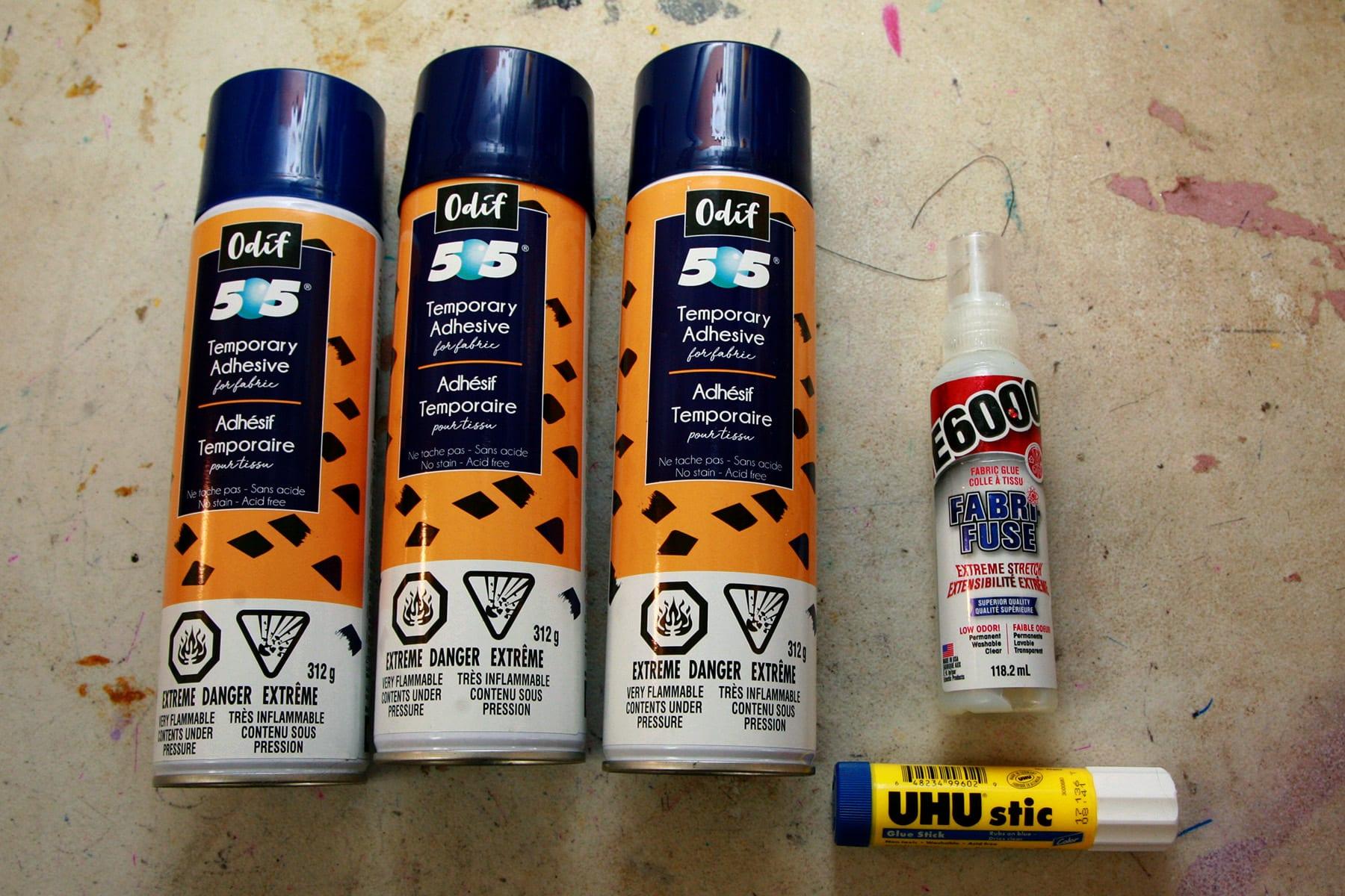 3 bottles of 505 spray adhesive are laid out next to a bottle of E-6000 Fabri-Fuse, and a glue stick.