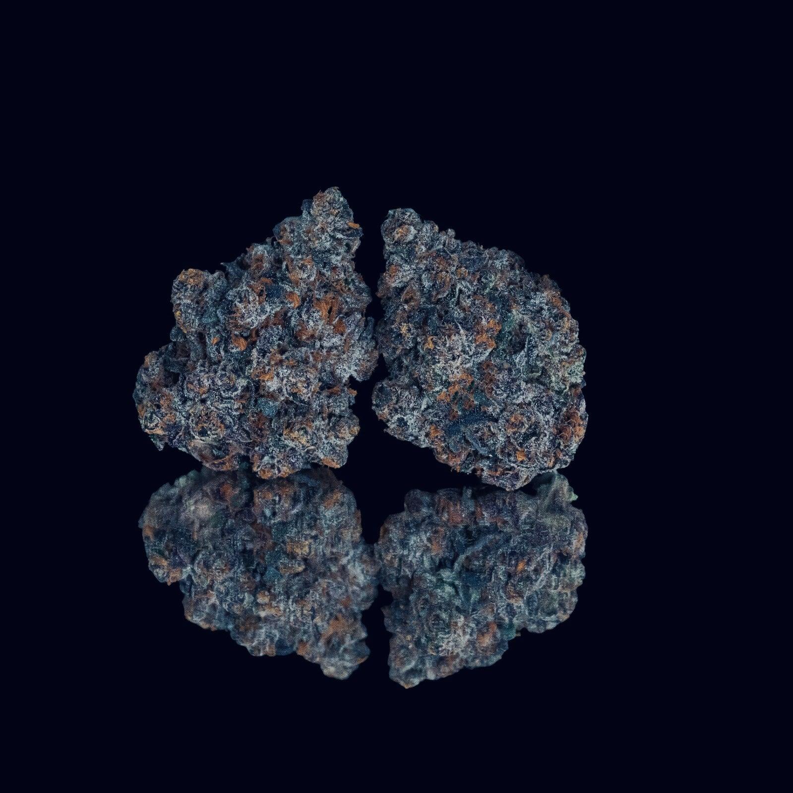 Two nugs of cannabis flower from the Grape Ape strain rest against each other above their reflection on a black surface.