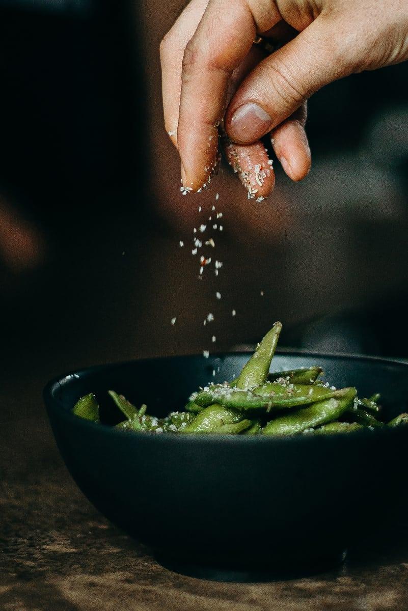 A hand sprinkling coarse salt over a bowl of edamame in the pod