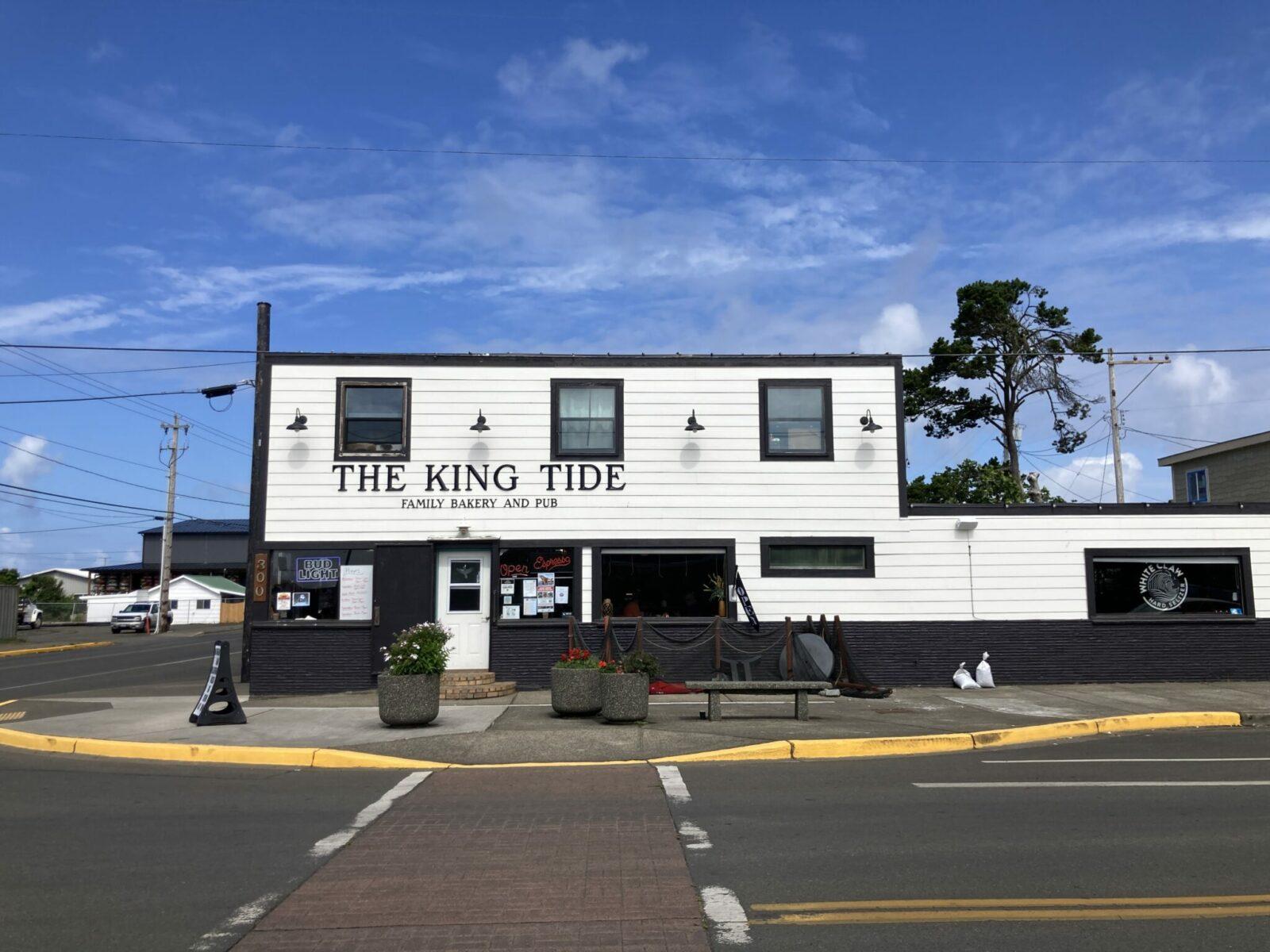 A white painted wooden building with black trim on a street corner. The sign says King Tide Family bakery and pub