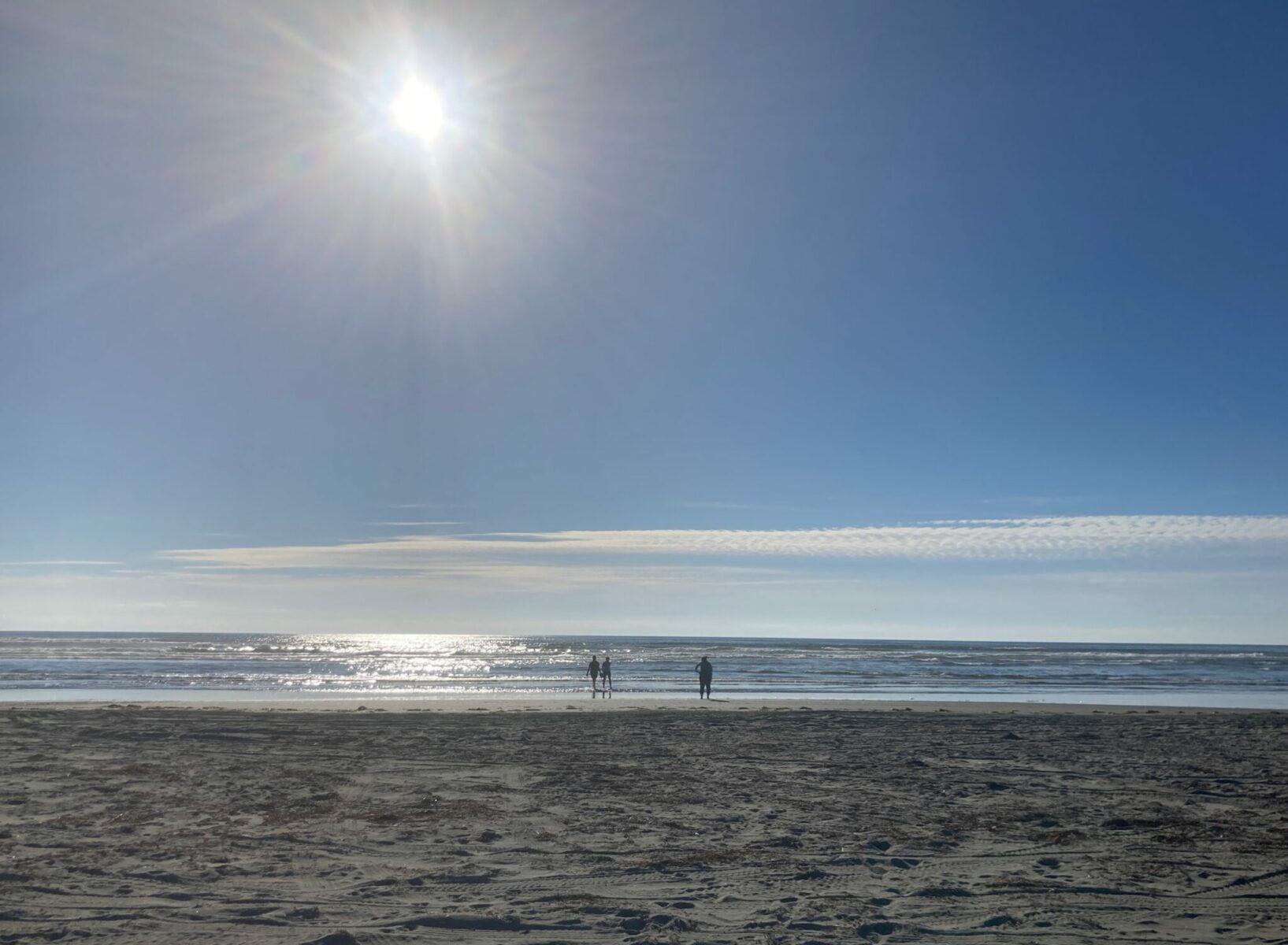 The sun getting low in the sky but still bright over the Pacific Ocean at a sandy beach. Three people are silouetted in the distance at the edge of the water