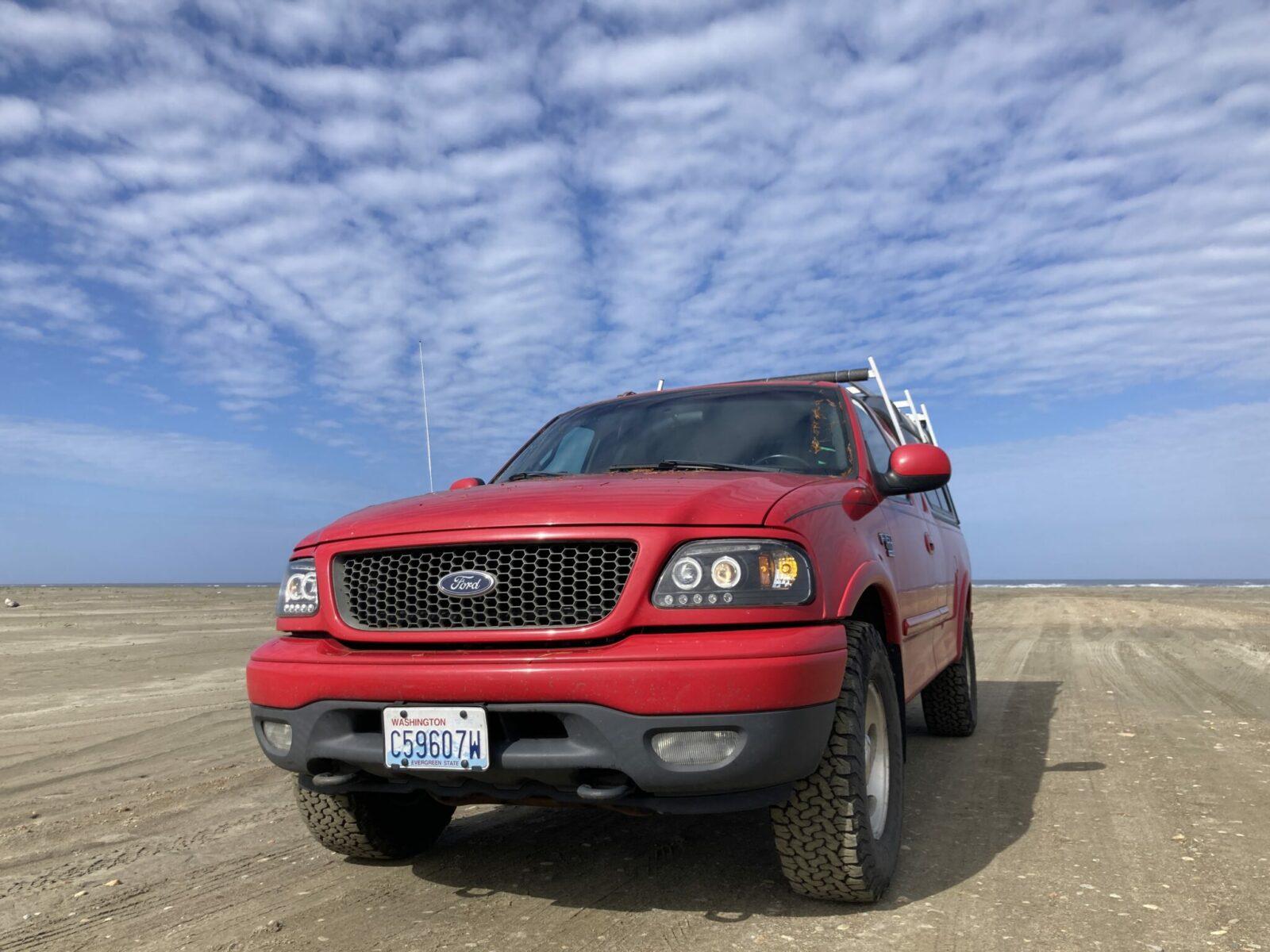 A close up of a bright red pickup truck on the sand at the beach on a sunny day.