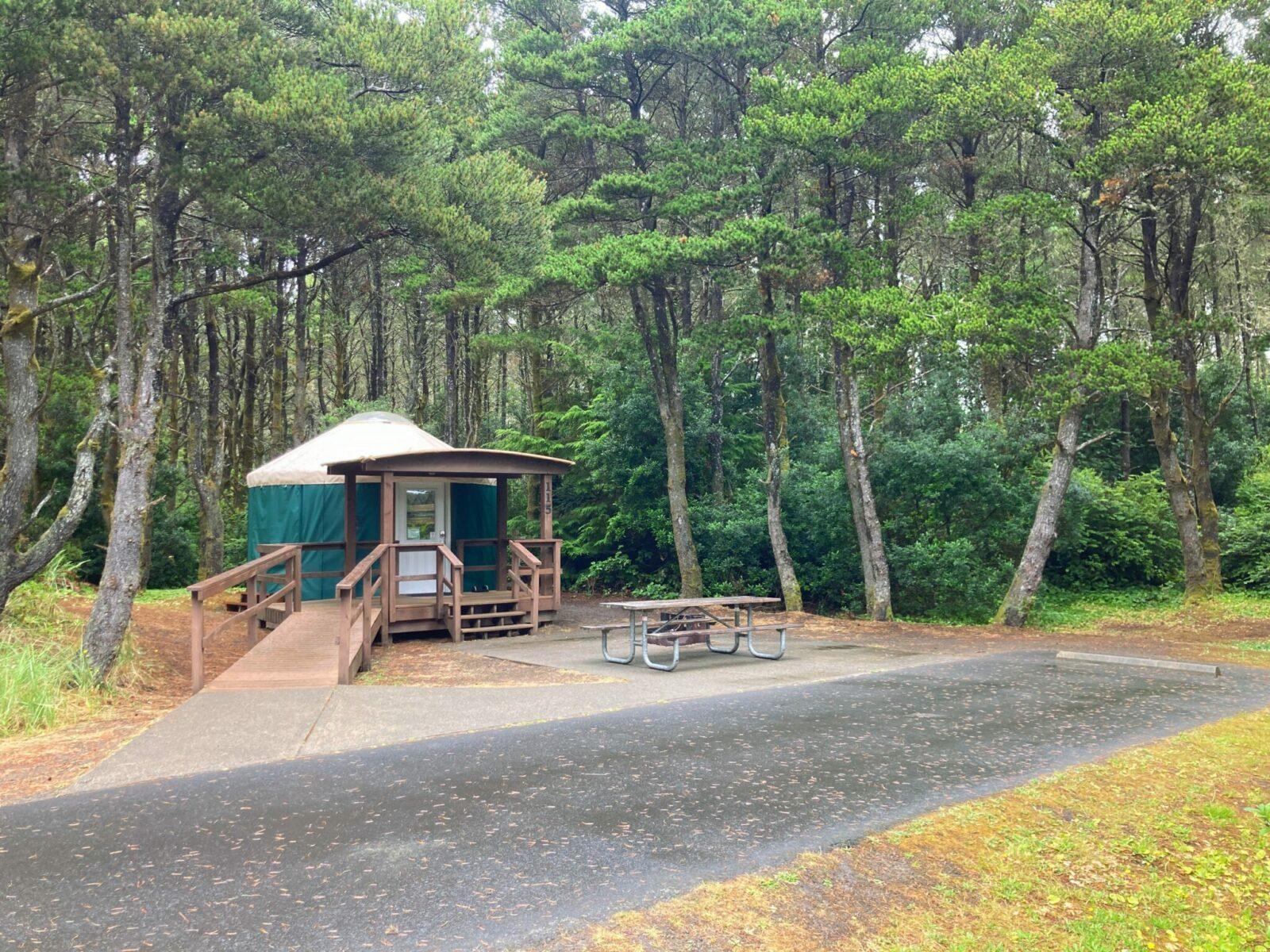 A green yurt with a tan roof surrounded by a wooden porch with stairs and a ramp. There is also a picnic table, firepit and paved parking spot. The yurt is surrounded by forest.