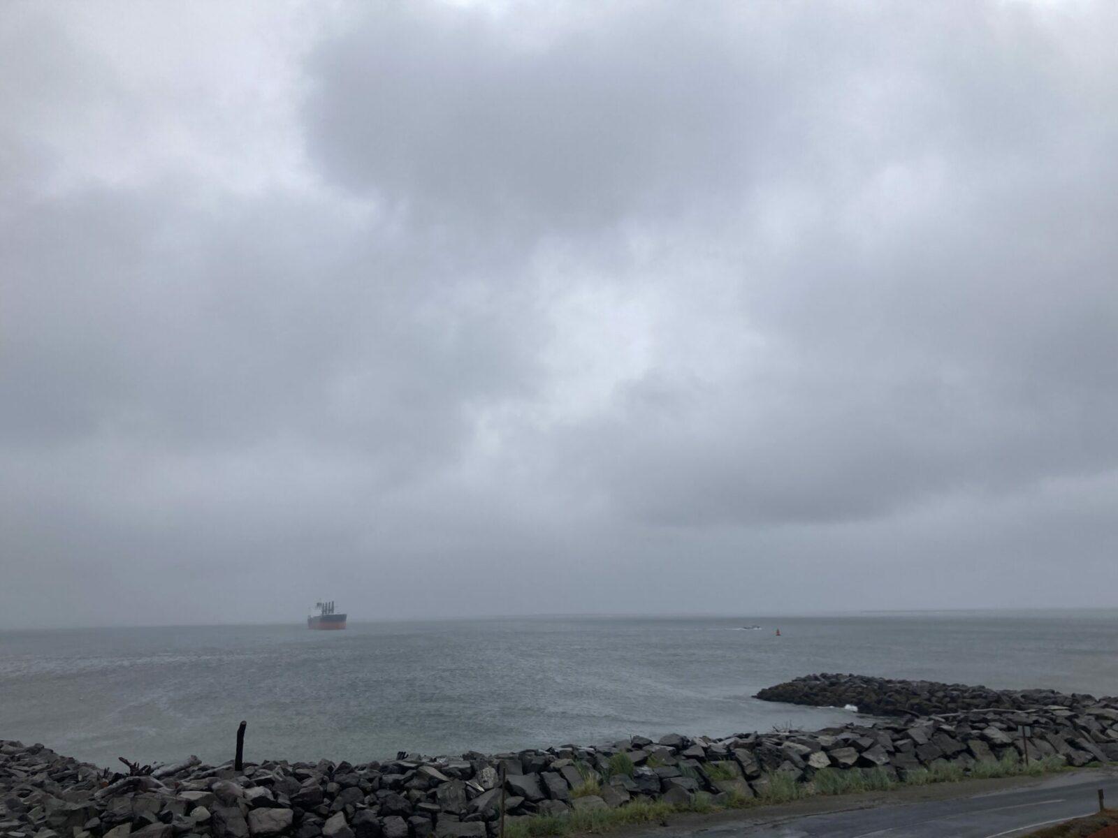A jetty and gray water with gray sky and a container ship just off shore