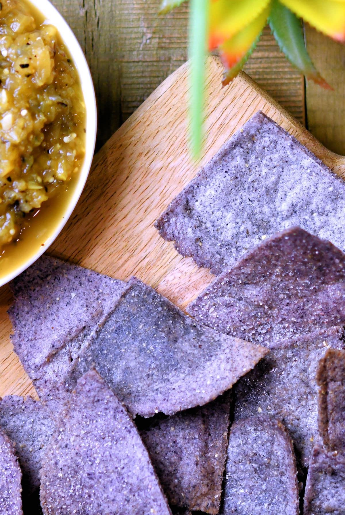 Homemade blue tortilla chips on a cutting board next to a bowl of salsa verde.