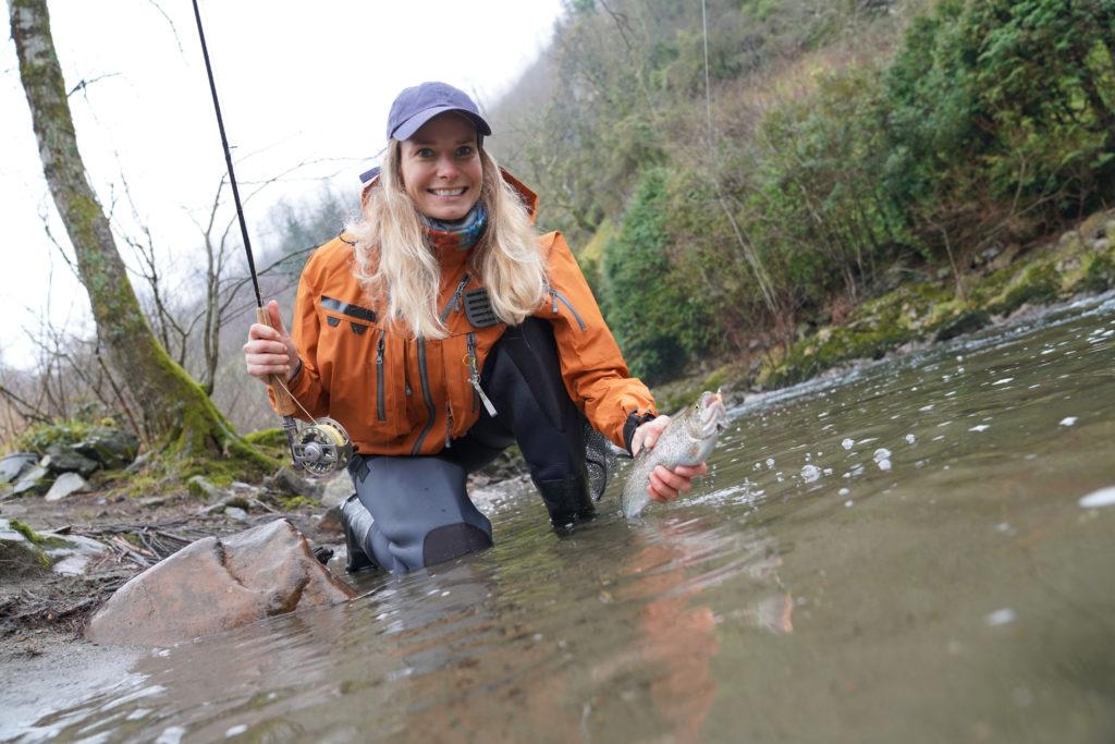 A blond fisherwoman kneeling in a river, holding her catch, a Rainbow Trout, with autumn landscape in the background
