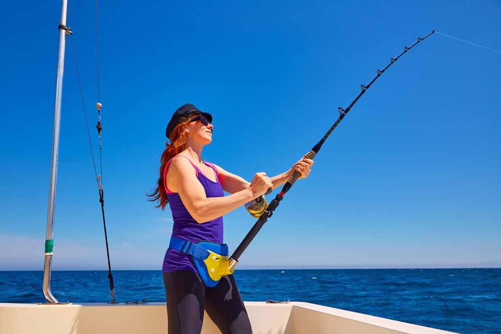A woman standing on a boat, in a cap and sunglasses, holding a fishing rod and battling fish, with blue skies and water in the background