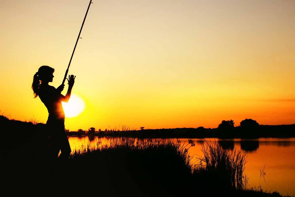 A silhouette of a woman angler reeling in her catch with sunset in the background