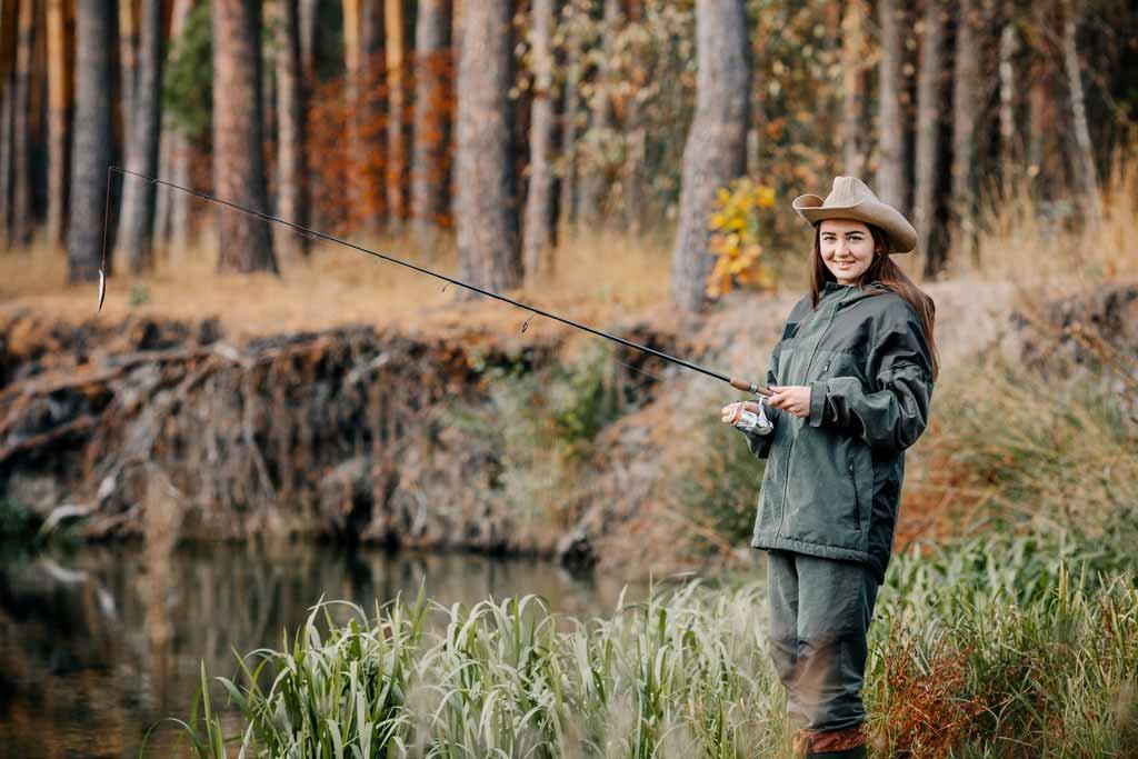 A woman in a hat and fishing clothes holding a rod with woods and foliage in the background