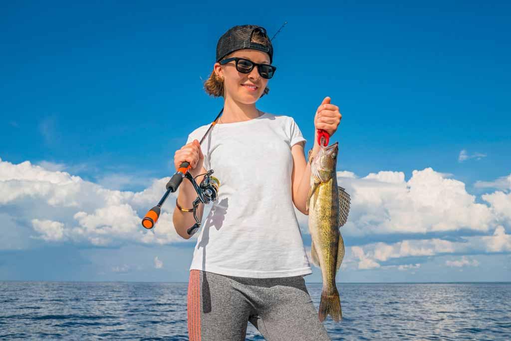 A young woman angler in a baselayer shirt, cap, and sunglasses holding a small Walleye