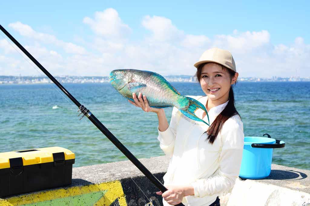 A young woman in a cap and long-sleeved shirt holding a fish with water and blue skies in the background