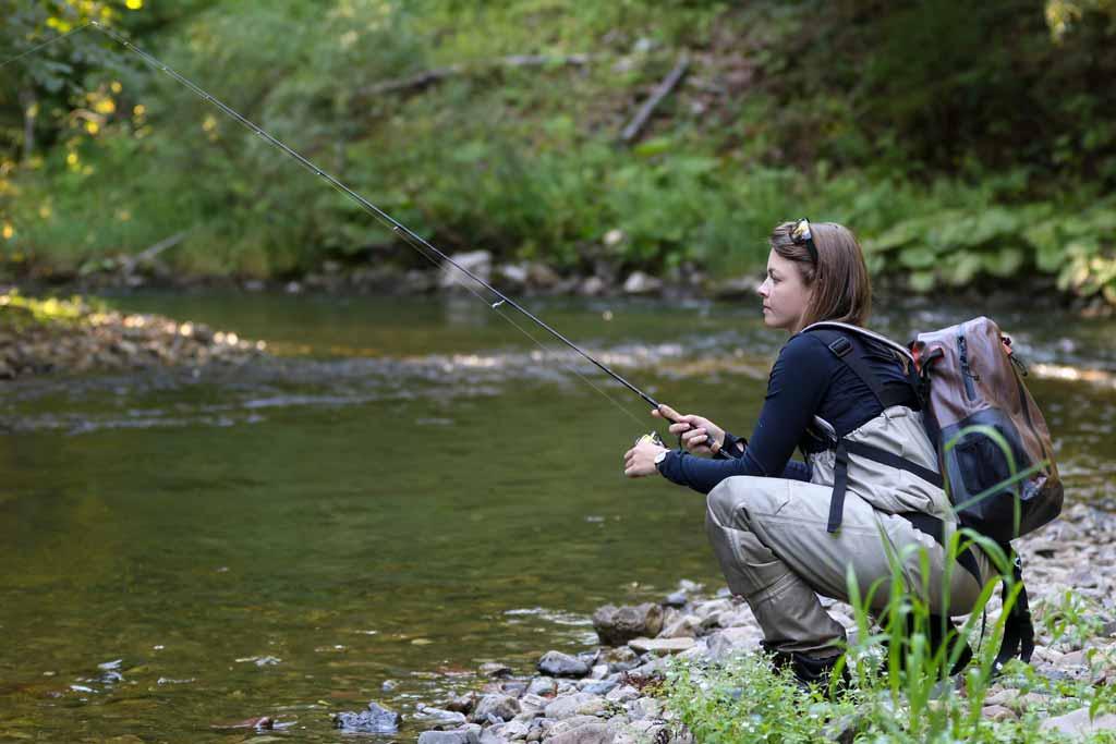 A woman angler squatting next to a river, holding a fishing rod