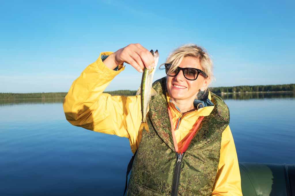 A female angler in a fishing vest and polarized sunglasses holding small Smelt with blue skies and water in the background
