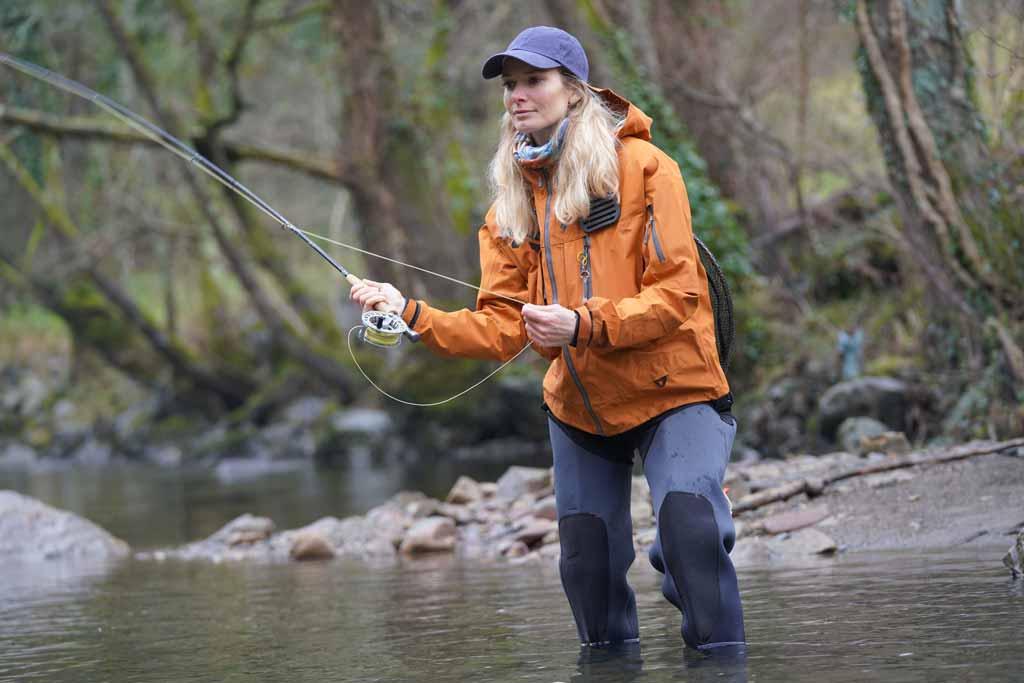 A woman angler fly fishing in knee deep water, with woods in the background