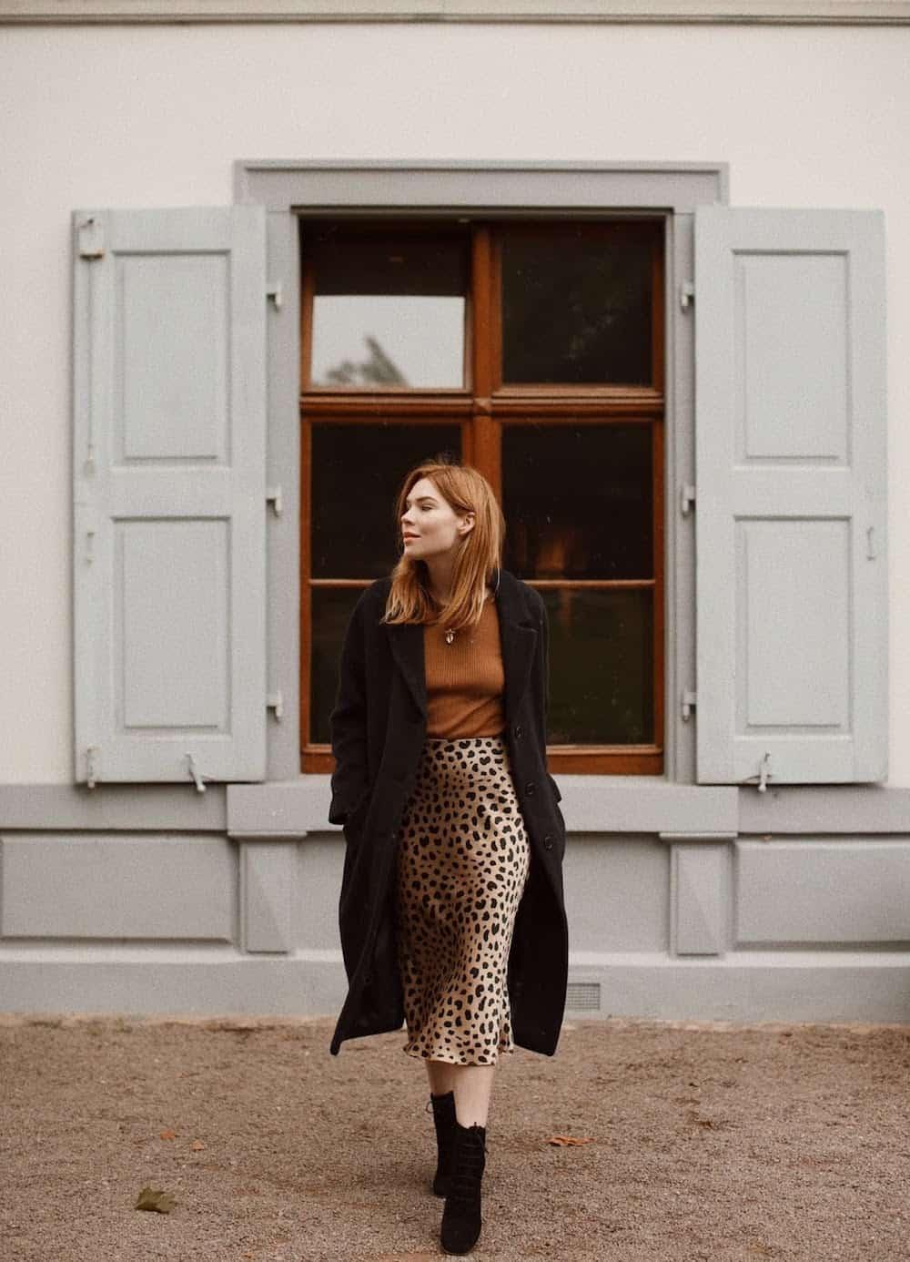 Woman wearing an animal print midi skirt, tan sweater, black booties and a black coat.