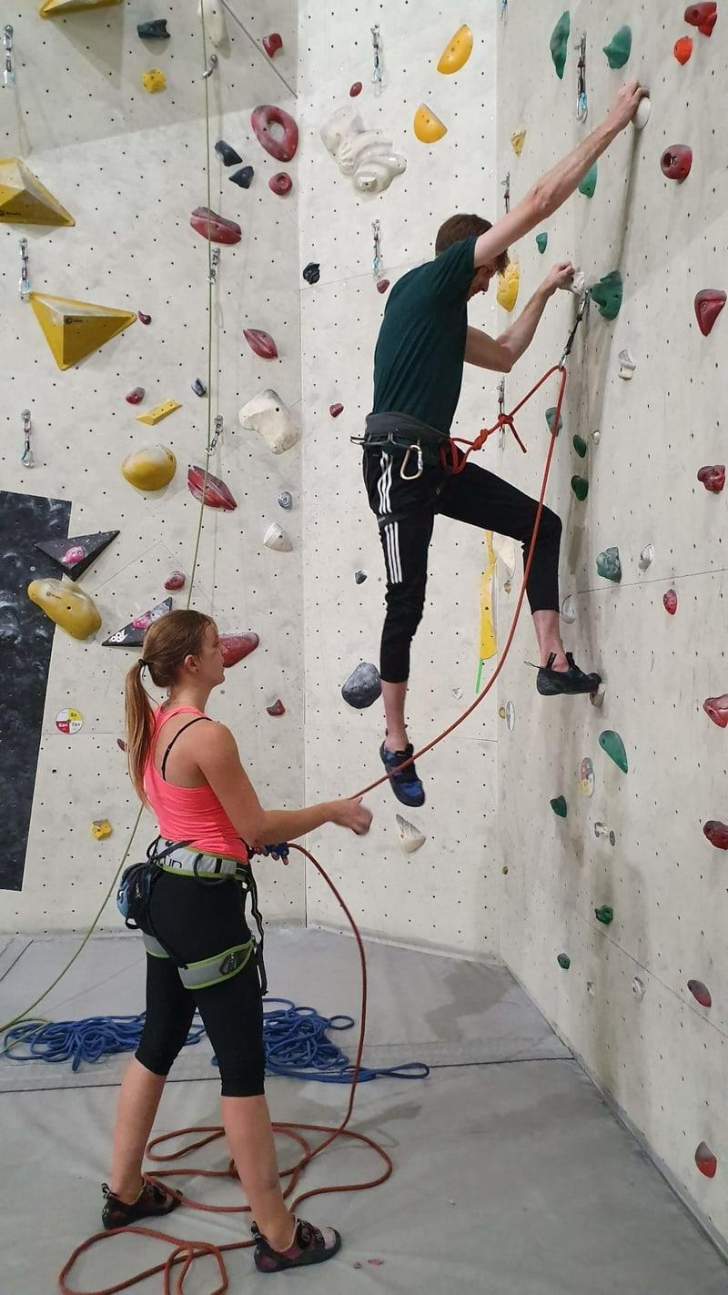 Two people rock climbing in an indoor wall, wearing tracksuits and tshirts