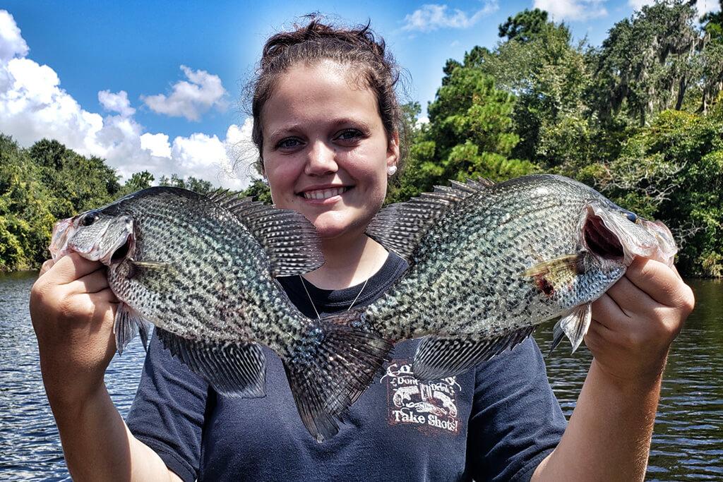 A young woman holding two large Black Crappie with a lake behind her