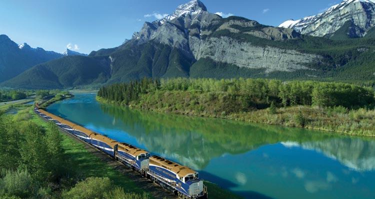 A train moves alongside a clear blue river below tree-covered mountains.