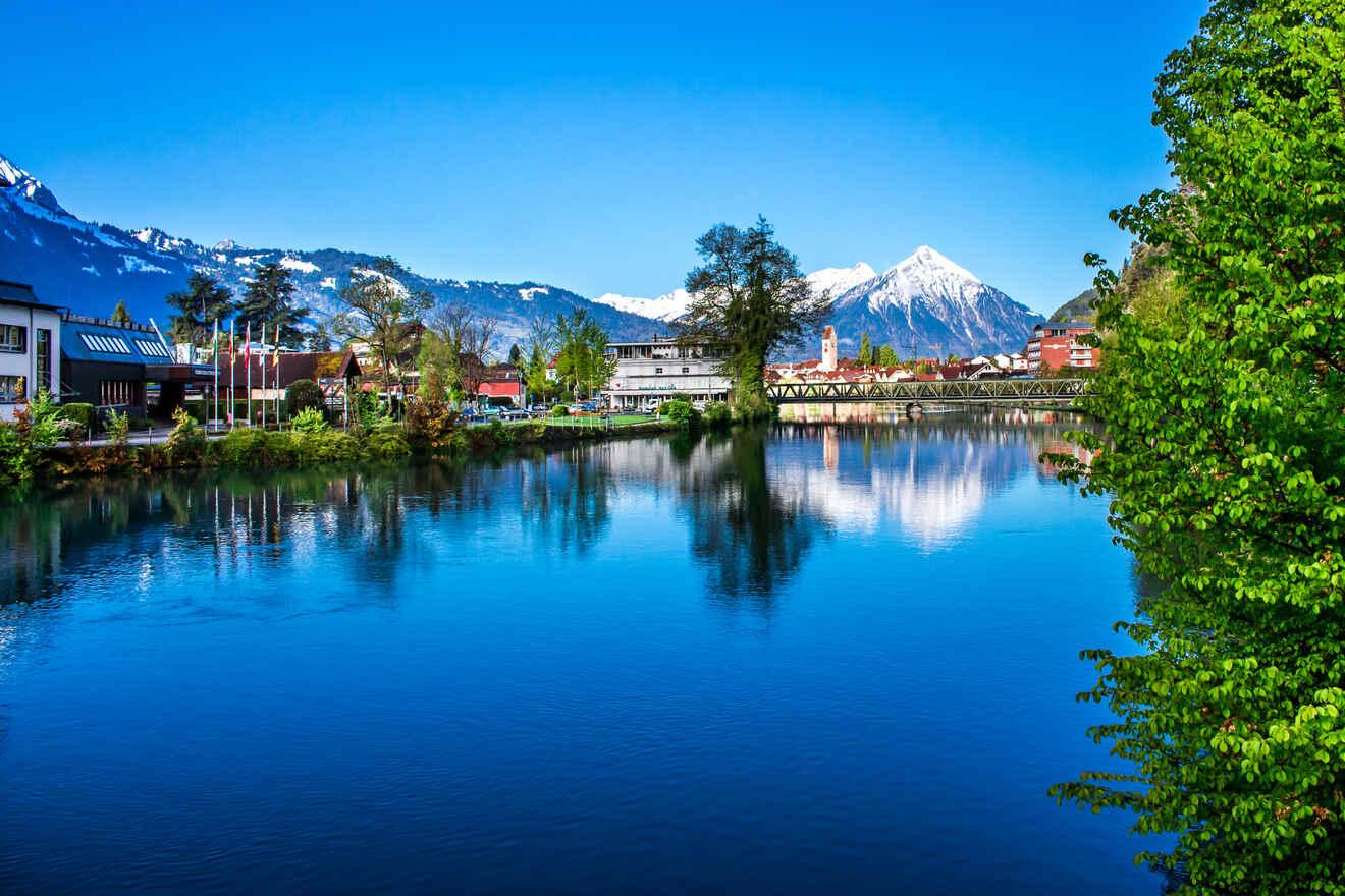 Serene riverside view in Interlaken with clear blue waters reflecting the surrounding greenery and snow-capped mountains in the distance under a blue sky