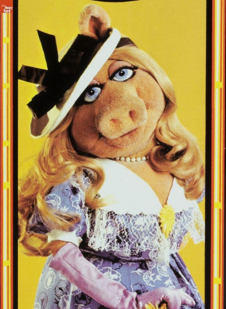 Pig puppet with blonde wavy hair wearing white hat with black ribbon, elbow-length purple gloves, and a purple dress with white collar