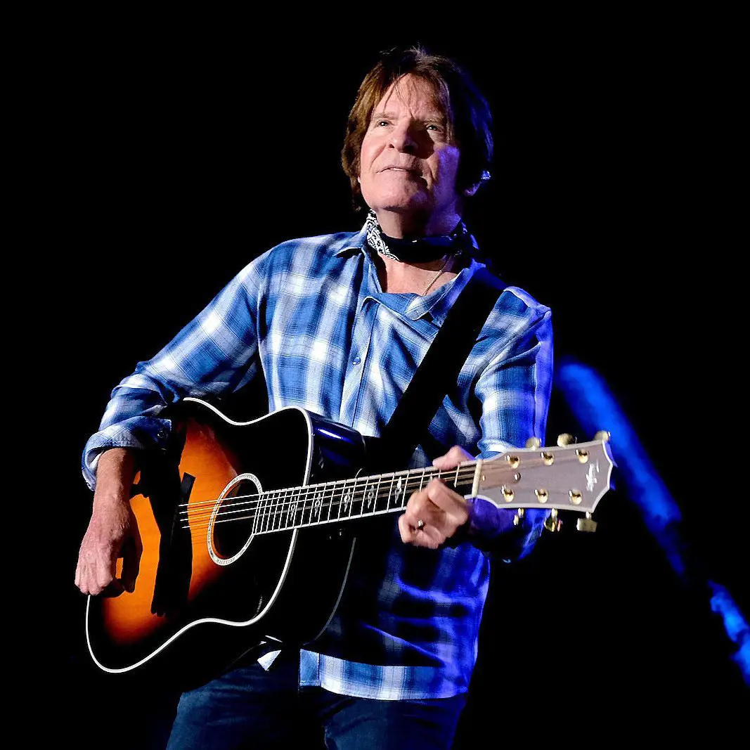 John Fogerty with a guitar and classic blue check shirt, promoting his tour in Friant, West Springfield, and Bridgeport