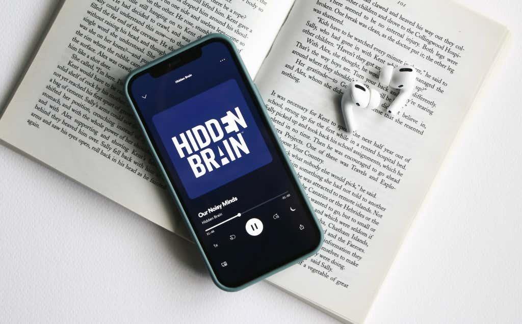An iPhone (with a podcast episode playing in Spotify) and AirPods placed on an open novel/book