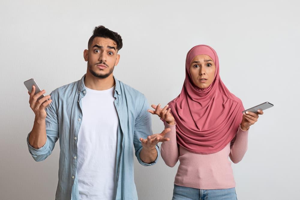 Confused muslim couple with smartphones looking at camera with frustration
