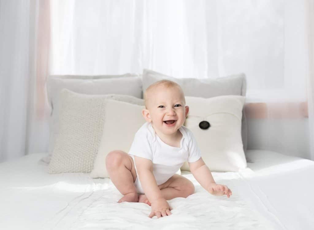 Do autistic babies laugh when tickled? A baby sits on a white bed, wearing a white onesie, and laughs at the camera.
