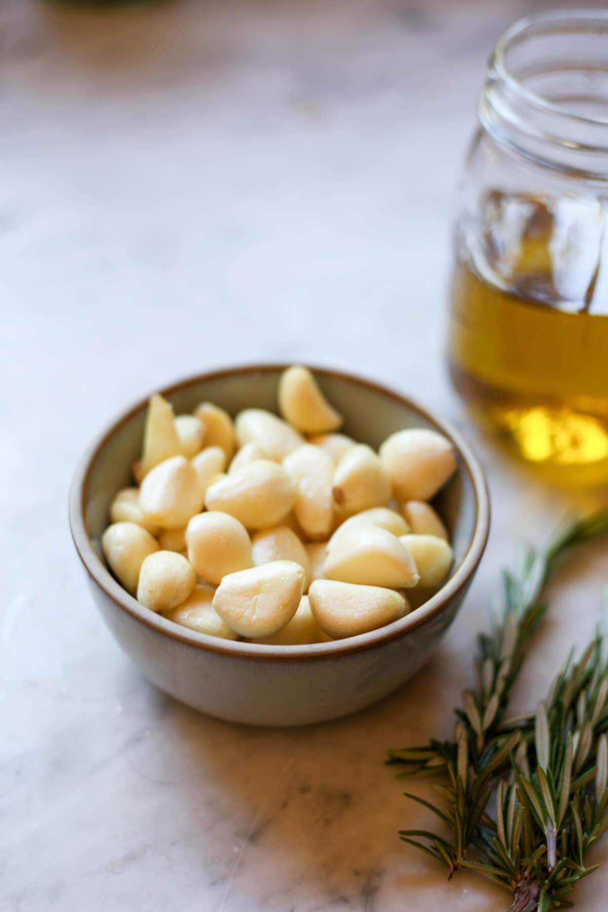 A small grey ceramic bowl filled with peeled garlic cloves on a marble counter with olive oil in the background and rosemary in the foreground.