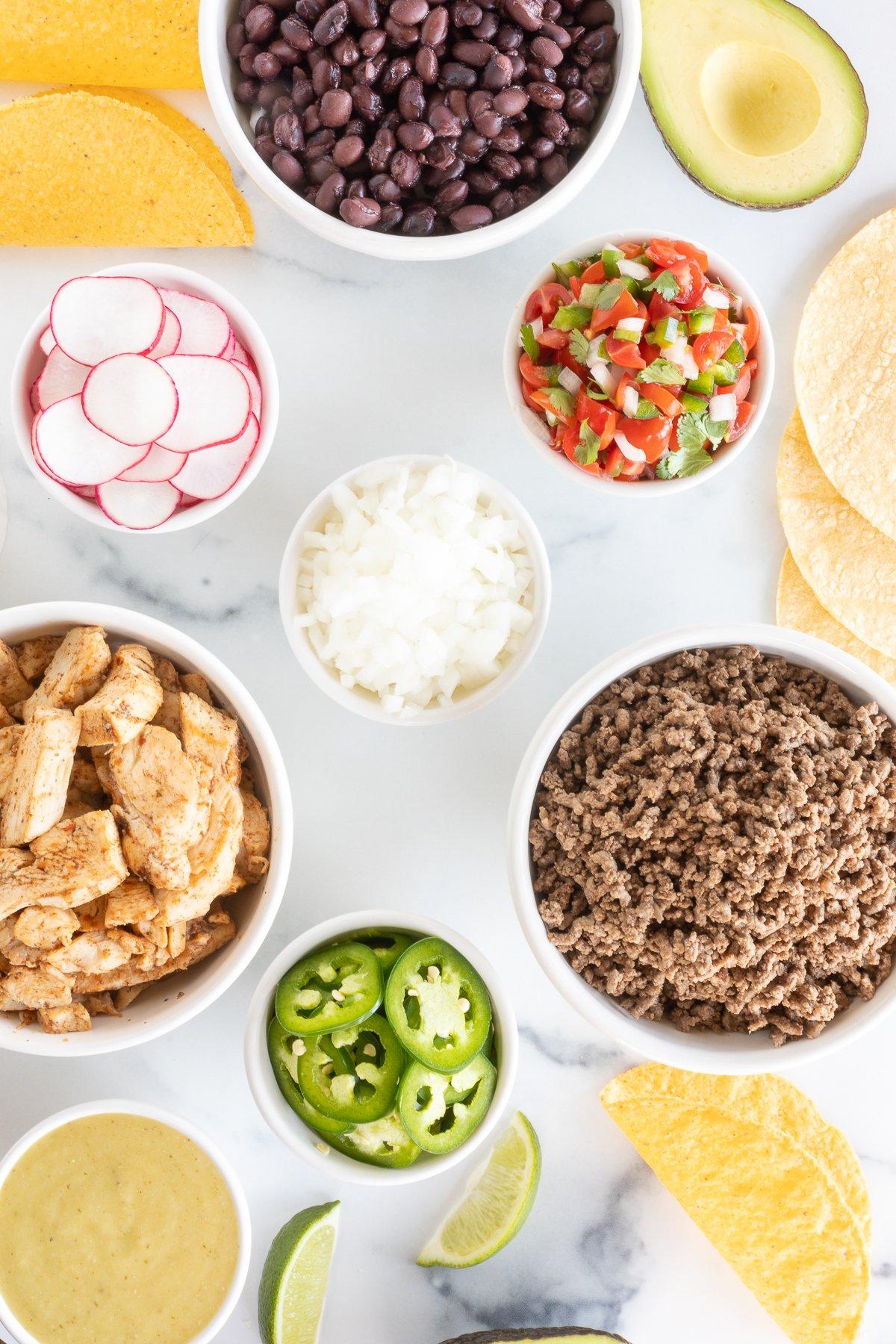 Taco toppings, including sliced radishes, chopped onions and sliced jalapenos on a marble countertop.