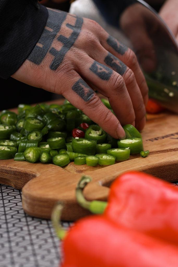 A Tattooed Person Holding a Green Chili Peppers on a Wooden Chopping Board