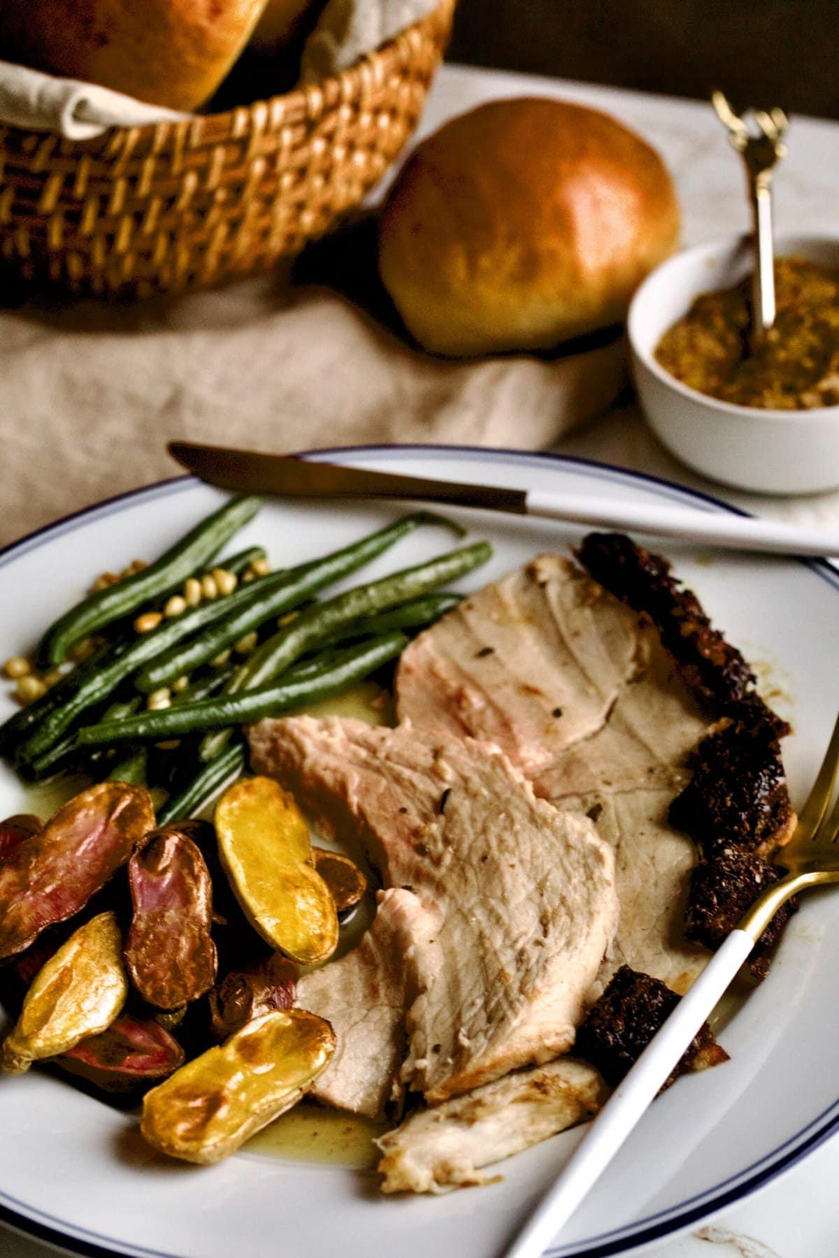 resh ham roast on a plate with roasted potatoes and green beans. A bread basket with rolls to the side.