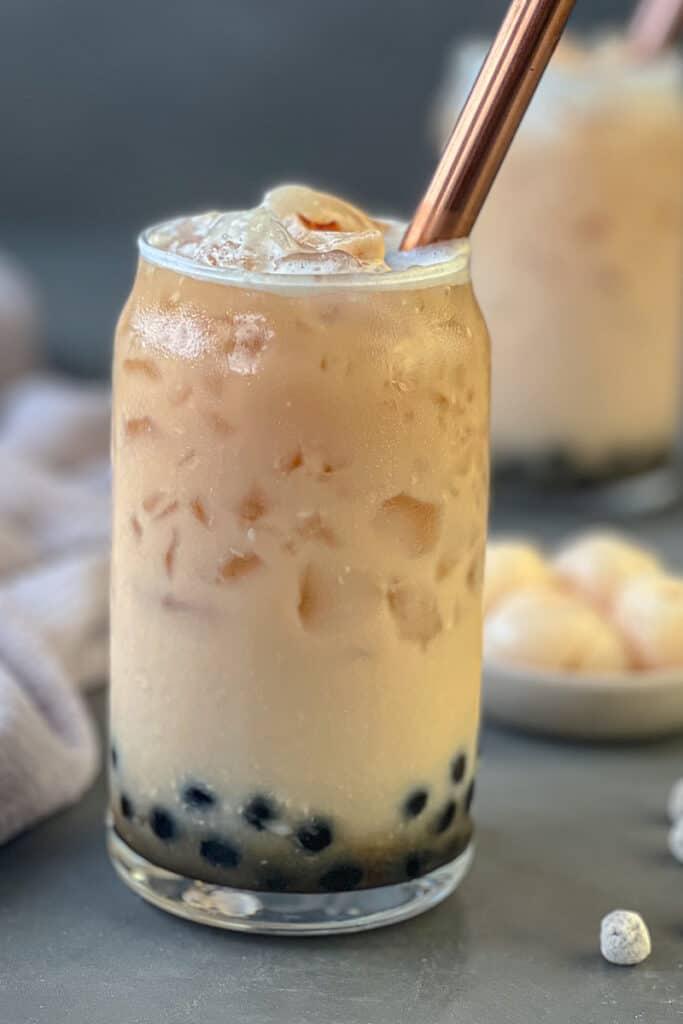 A tall glass of lychee bubble tea next to whole lychees and boba pearls.