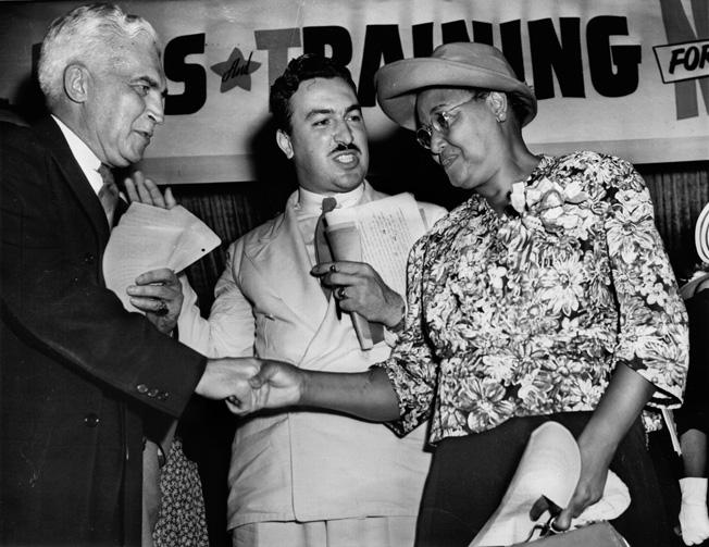 Paul V. McNutt, chairman of the War Manpower commission, shakes hands with Mrs. Connery Miller, mother of Dorie Miller. Reverend Adam Clayton Powell, Jr., looks on during a rally of the Negro Labor Victory Committee held in Harlem.