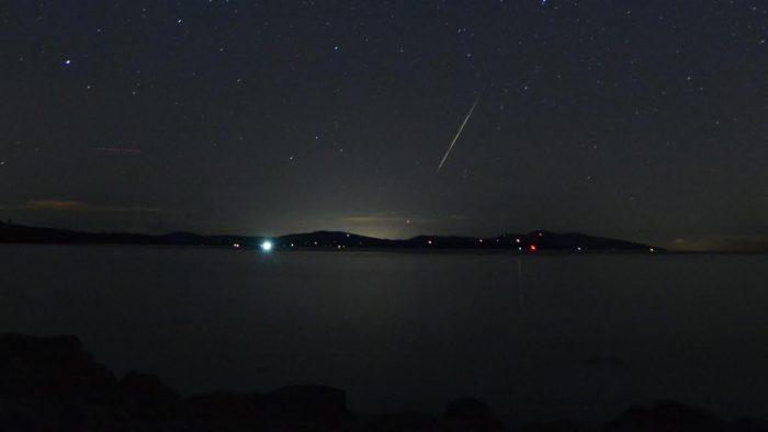 Meteor streak over low-lying hills with sea in foreground.